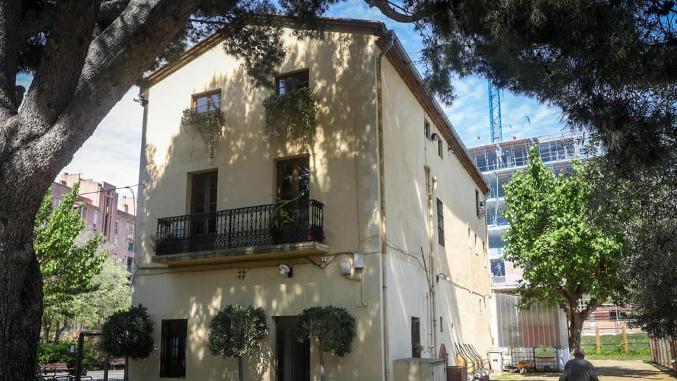 An old Catalan farmhouse (masía) with stone walls and a traditional tiled roof, surrounded by modern urban buildings in Barce
