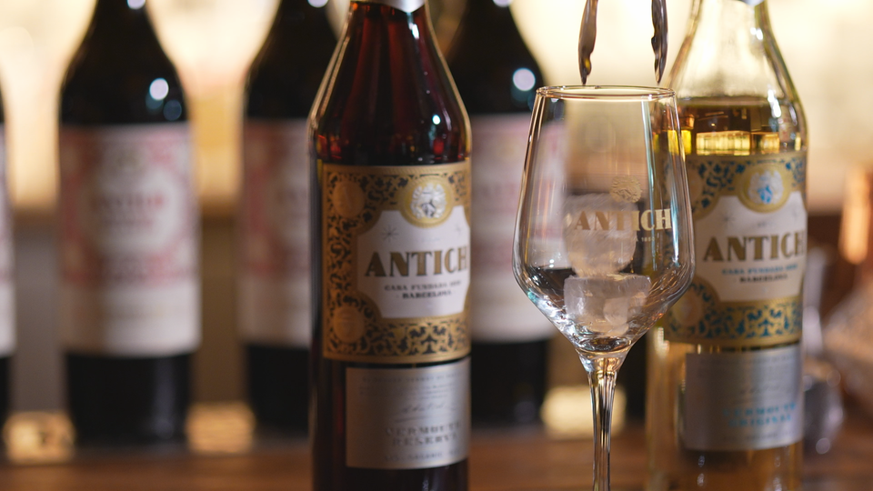 A bottle of Antich vermouth with a classic label, set against a blurred background of a traditional Barcelona bar interior.
