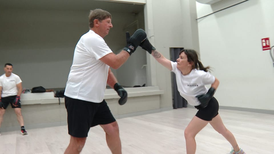 Two men in traditional martial arts attire demonstrate 'joc maonès' moves indoors, one in a defensive stance, the other attac