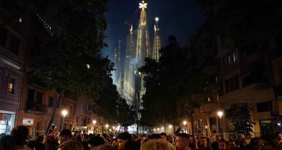 The Sagrada Família's Jesus Christ Tower illuminated at night, topped with a large cross, with crowds gathered below.