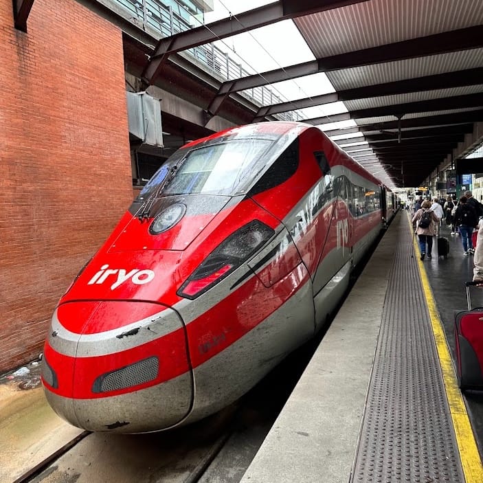 A red and grey Iryo high-speed train is parked at a station platform. Passengers with luggage are visible further down the platform.