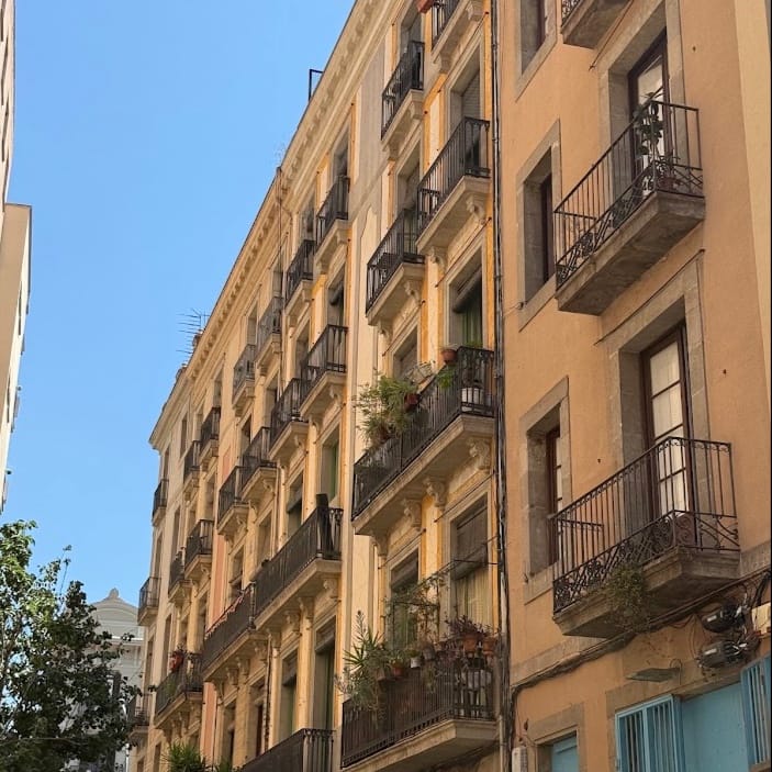 A low-angle view of several tall, old residential buildings with numerous balconies, some adorned with potted plants, under a clear blue sky.