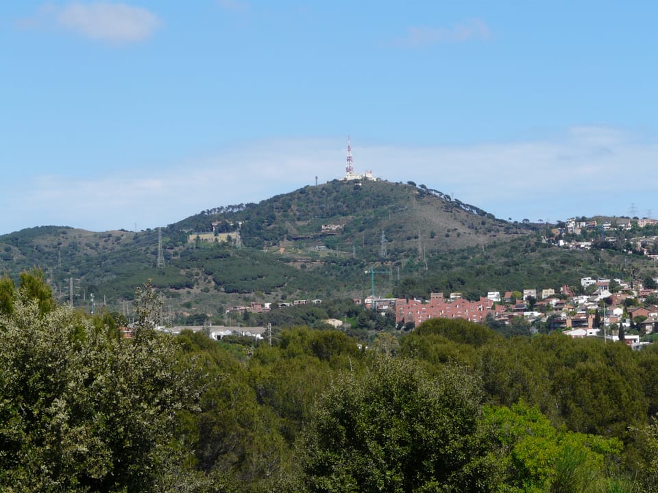Smoke rising from the Collserola mountains near Barcelona, with emergency vehicles and a Guardia Urbana car blocking access to a forest road.