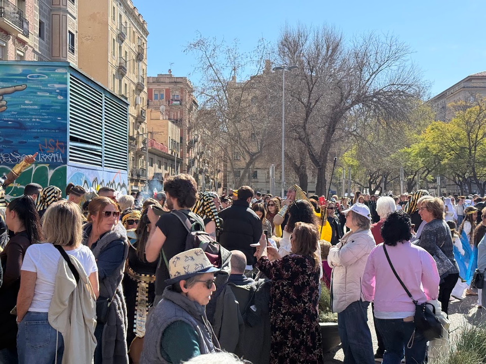 A large crowd of people fills a city street on a sunny day, with buildings and bare trees in the background.