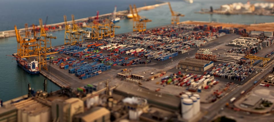 Aerial view of the Port of Barcelona with cargo ships, container stacks, and vehicle storage lots visible along the waterfront.