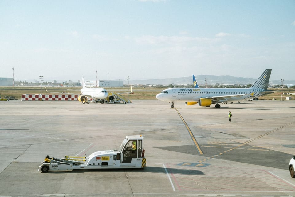 A Vueling aircraft taxiing at Barcelona El Prat Airport under a clear sky, with terminal buildings in the background.