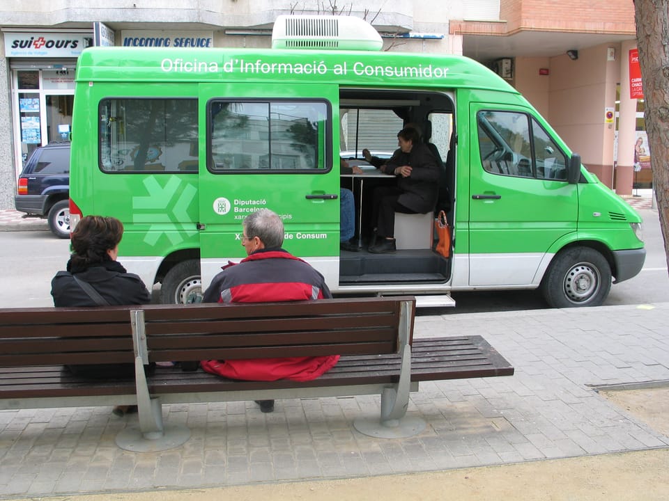 A digital illustration showing a person using a tablet to connect via video call with a consumer advisor, set against a backdrop of small towns in the Barcelona province.