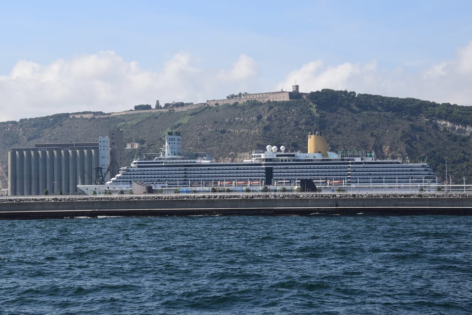 A cruise ship docked at the Port of Barcelona near Pont d'Europa, with city skyline in the background.