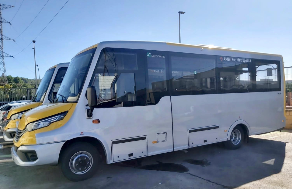 A modern AMB minibus on a quiet residential street in Molins de Rei, surrounded by green hills and low-rise housing.
