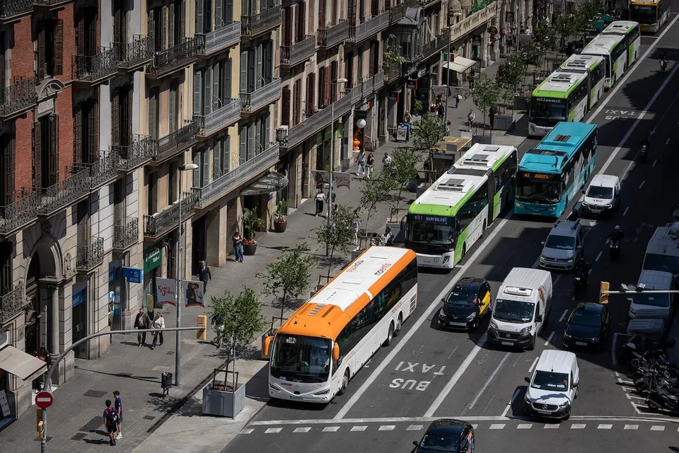 Intercity buses parked along a busy street in Barcelona's city centre, with pedestrians walking on the pavement and historic 