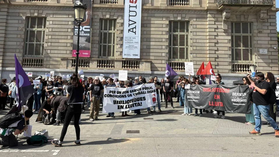 Library workers marching in a protest through Barcelona, holding banners and signs, with city buildings in the background.