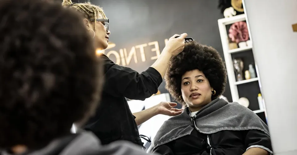 A woman with voluminous natural Afro hair smiling while a hairdresser styles it in a salon.