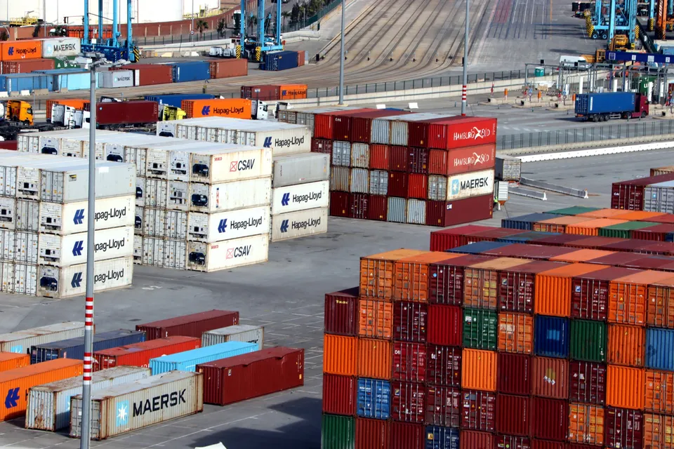 Aerial view of the Port of Barcelona with cargo ships, cranes, and container terminals under a clear sky.