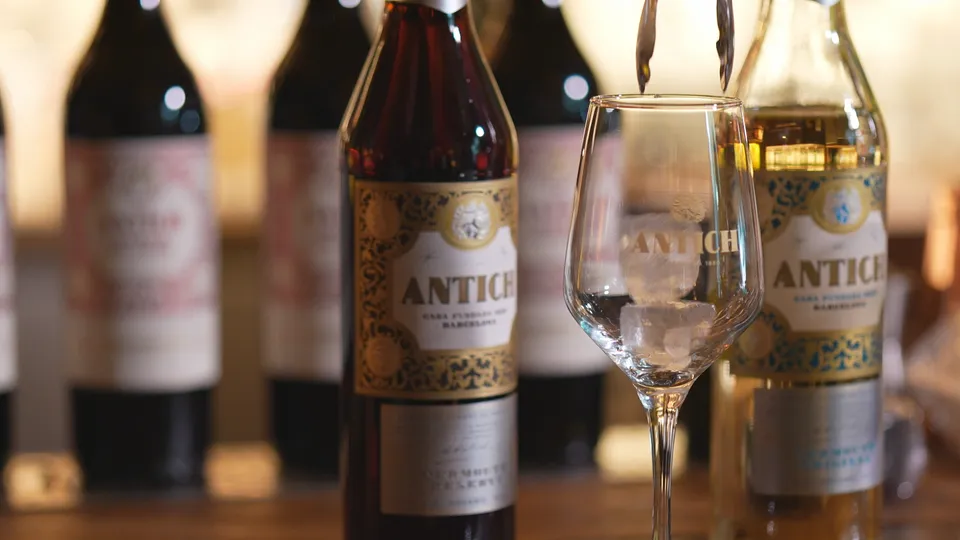 A bottle of Antich vermouth with a classic label, set against a blurred background of a traditional Barcelona bar interior.