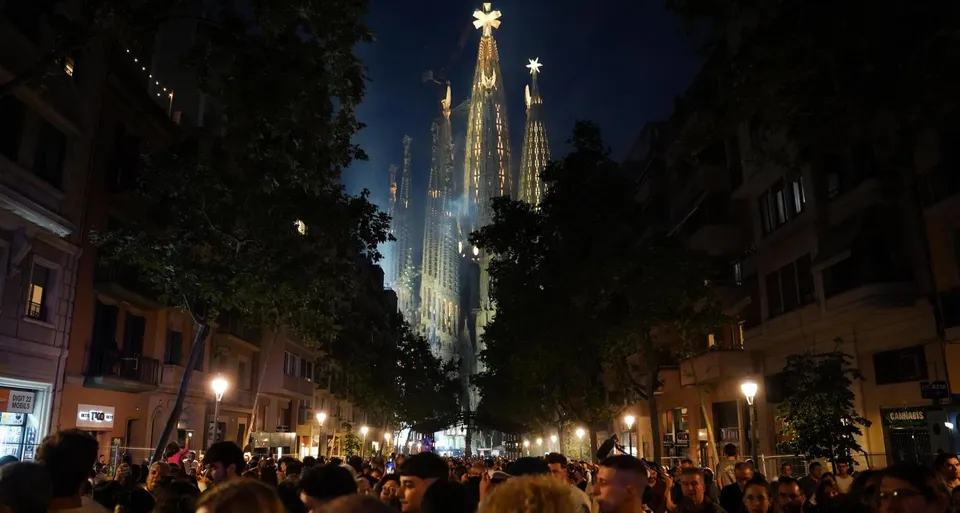 The illuminated Jesus Christ Tower of Sagrada Família with a large cross on top, against a night sky.