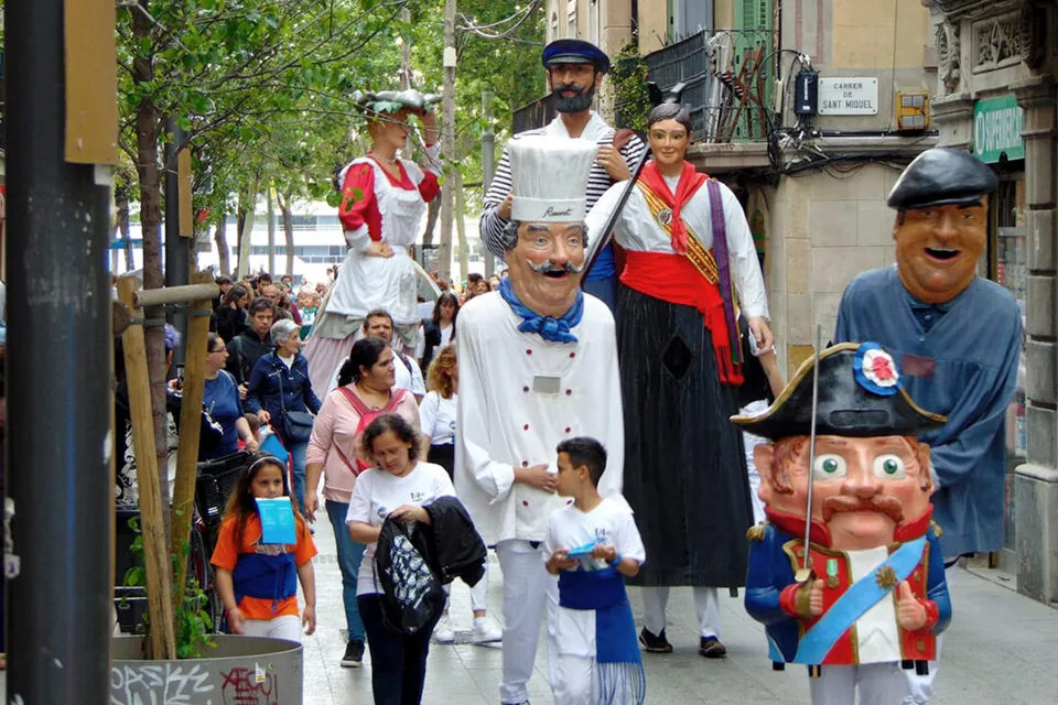 A festive street scene in Barceloneta during Festa Major, with decorated streets and a procession featuring traditional giants and local residents.